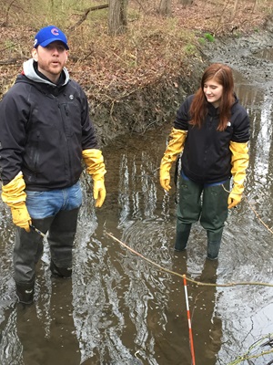 Trimby and one of his students retrieve a tray of tiles from a stream near their campus as part of an aquatic ecology lab in his Introductory Biology II class.
