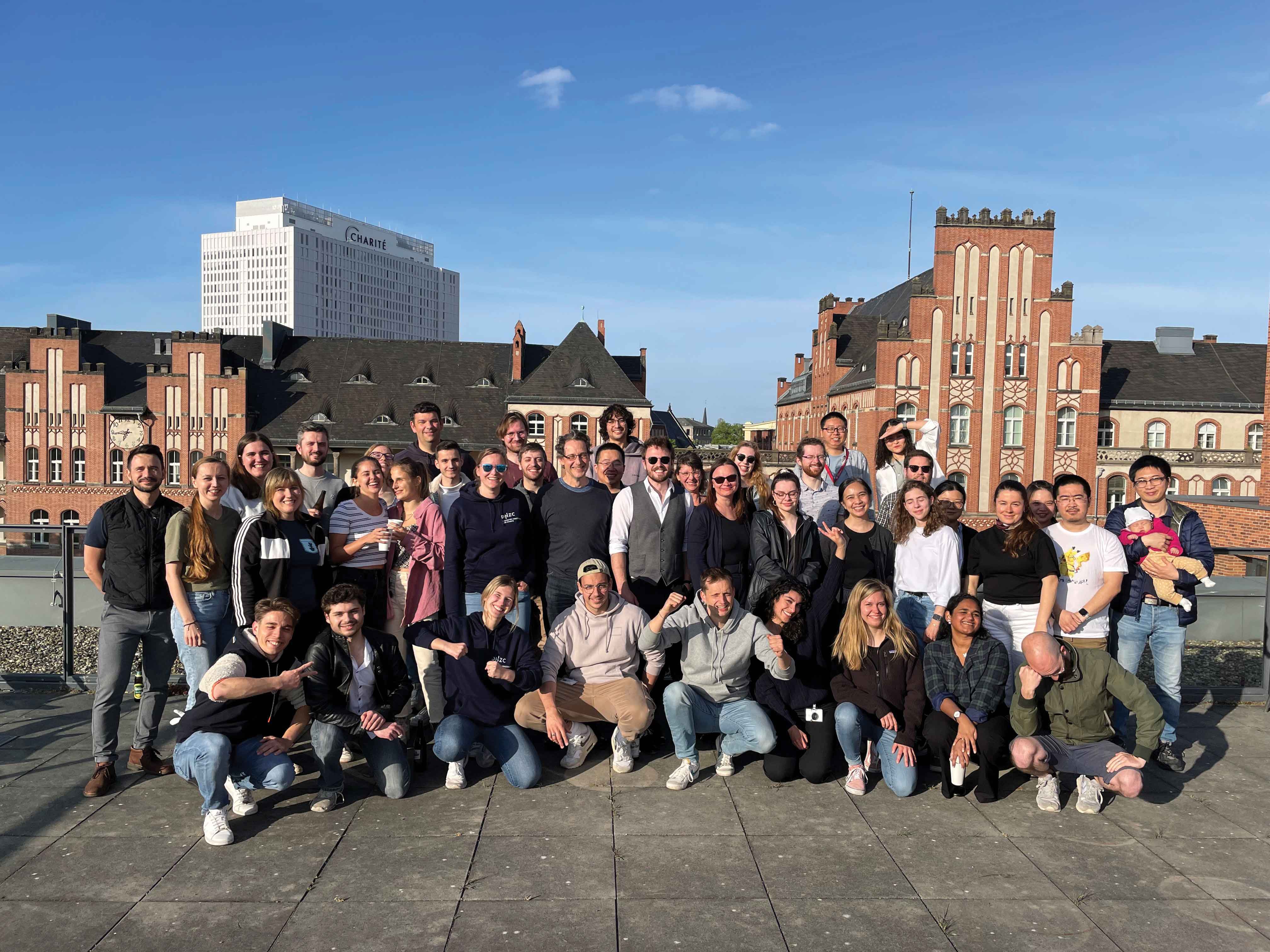 Feature-Art_Kuebler-Profile_photo-2 A large group posed assembled in three rows in front of some classic brick campus buildings